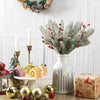 A white ceramic vase holds a bouquet of frosted pine needles and red berries on a wooden surface.