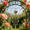 A gold heart-shaped window pendant with a faceted crystal charm hangs from an ornate metal arch covered in pink roses.