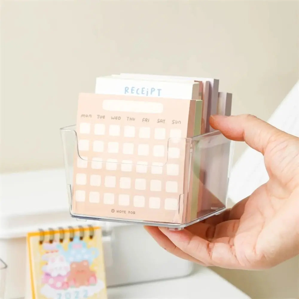 Une main tient un organiseur de bureau en acrylique transparent avec des blocs-notes empilés dans diverses couleurs pastel.
