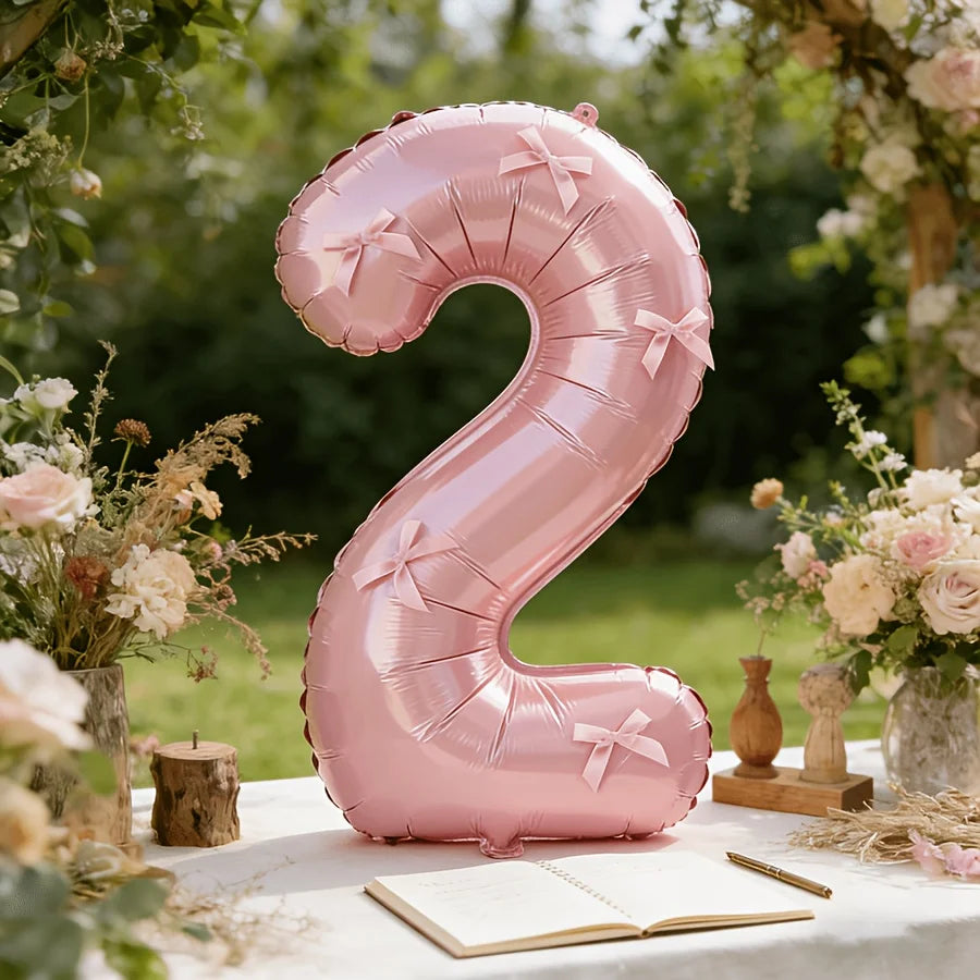 A large pink foil number balloon shaped like the number 2 with a bow, placed on a white table surrounded by floral greenery.
