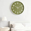 A minimalist green wall clock with white numbers and hands hanging above a white bed.