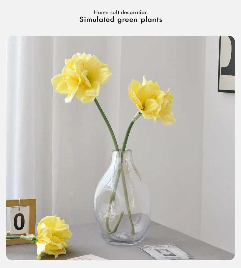 A yellow artificial clivia amaryllis flower in a clear glass vase on a light gray surface.