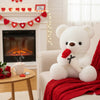 A white bear plush toy with a red rose, sitting on a red blanket next to a fireplace.