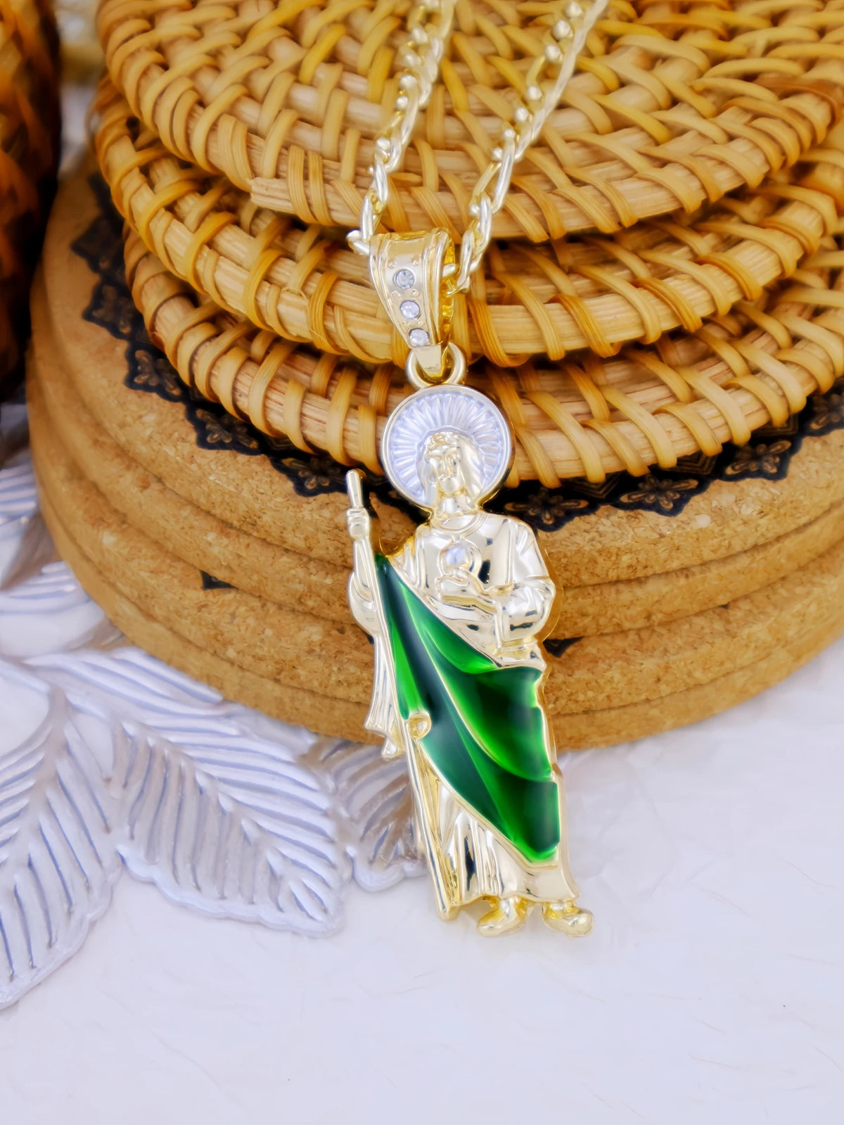 A gold-plated Saint Jude pendant necklace featuring a figure in green drapery and a halo, resting on woven mats.