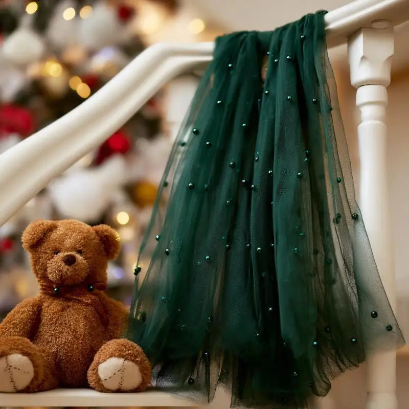 A dark green sheer fabric flag with a beaded trim is displayed next to a brown teddy bear on a white wooden railing.