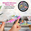 A hand is launching a pink dinosaur toy from a slingshot towards a target, with other dinosaur toys scattered nearby.