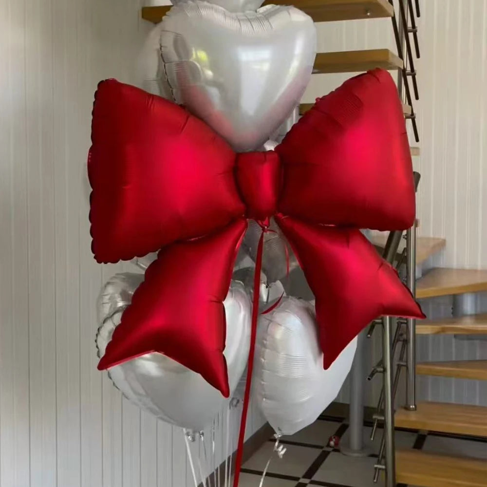 A red bow-shaped foil balloon is attached to a white heart balloon, displayed next to a wooden ladder.