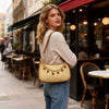 A young woman wearing a beige sweater and jeans holds a gold-toned jewelry bag with a woven texture and red stones in an outd