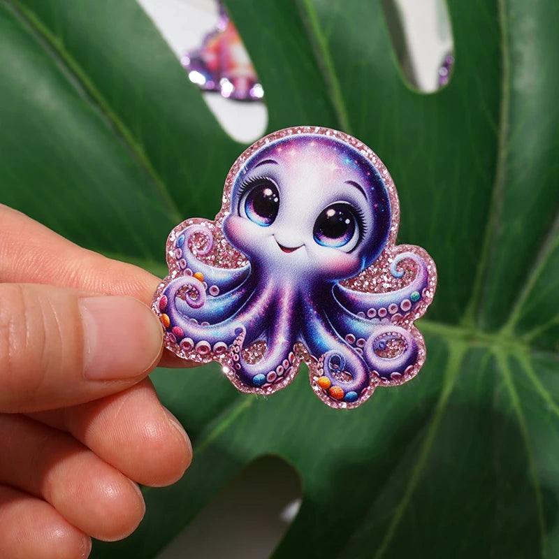 A pink glitter acrylic charm shaped like a purple octopus with large eyes, held by a hand against a green leaf background.