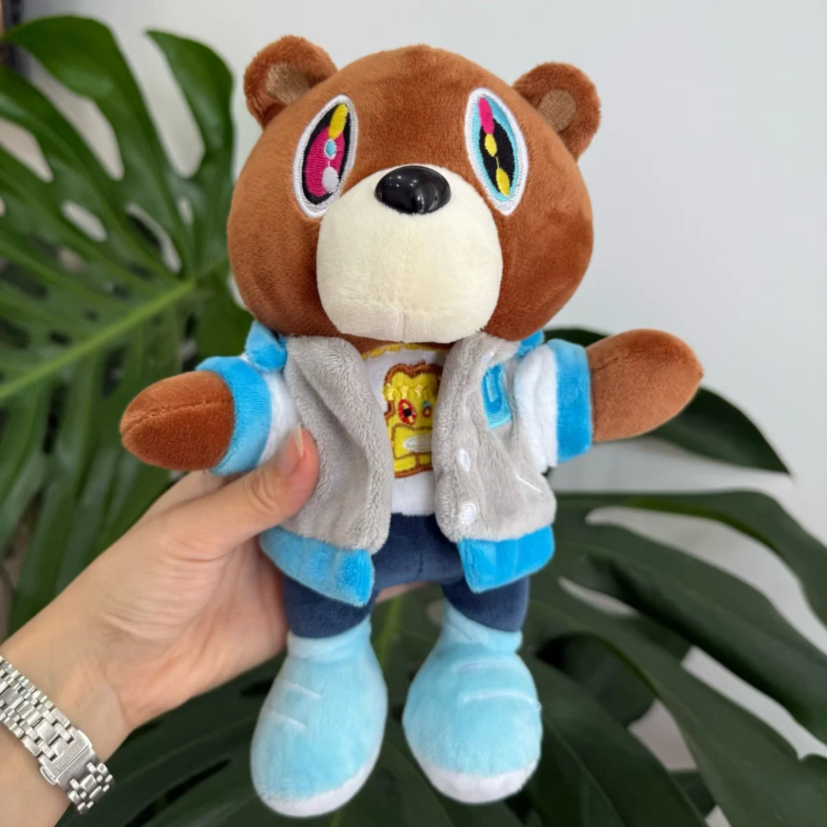 A brown bear plush toy with colorful eyes and a light gray jacket, held by a hand against a green leaf background.