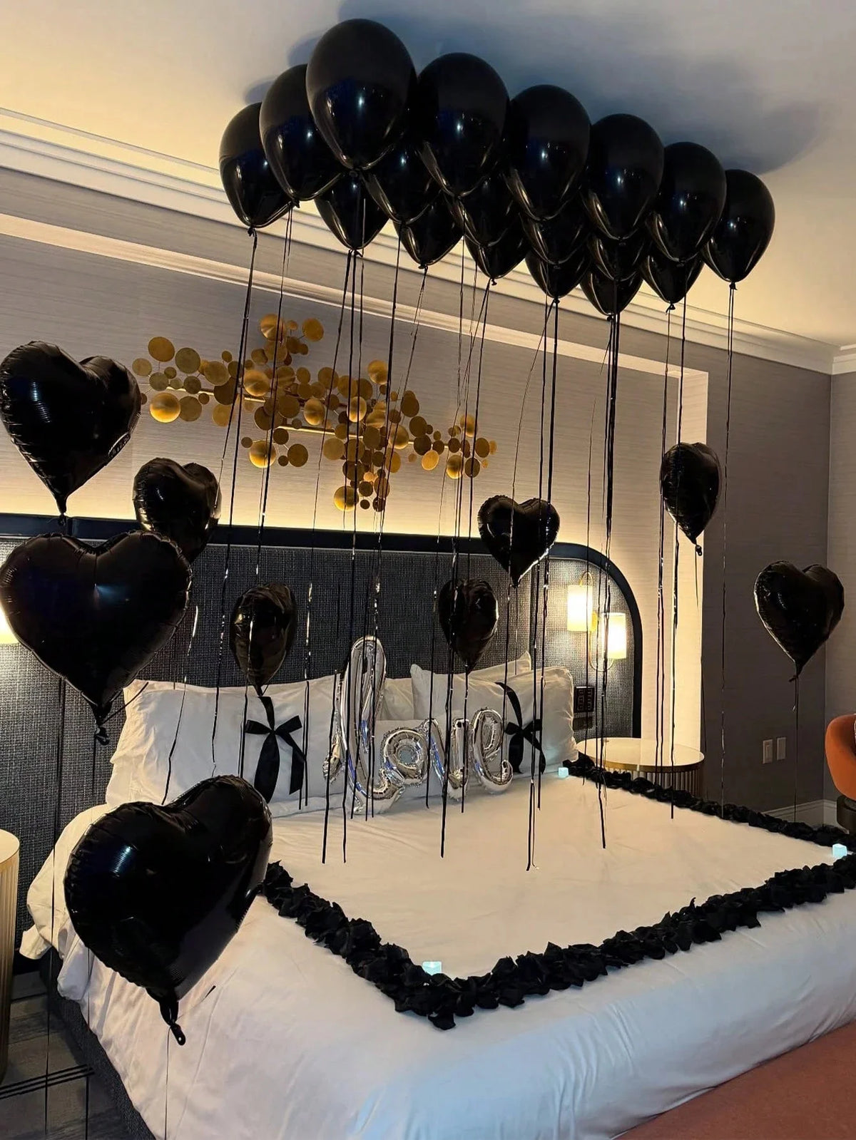 Black balloons with foil letters and rose petals hanging from the ceiling above a bed.
