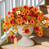 A vibrant arrangement of orange, red, and yellow artificial petunia bundles in a light gray pedestal vase on wooden steps.