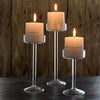 Three elegant tall glass candles with lit flames displayed on a dark wooden surface.