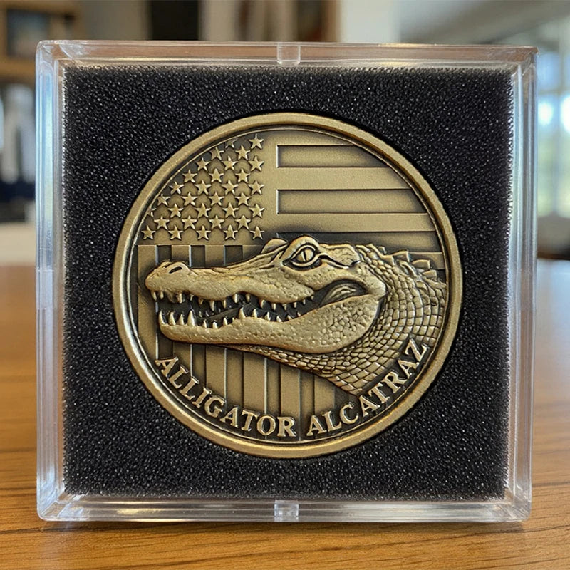 A clear plastic case holds a gold-colored coin featuring an alligator head design and the text Alligator Alcatraz