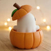 A white elf-like figure with an orange pumpkin top and green stem is displayed inside a carved orange pumpkin bowl.