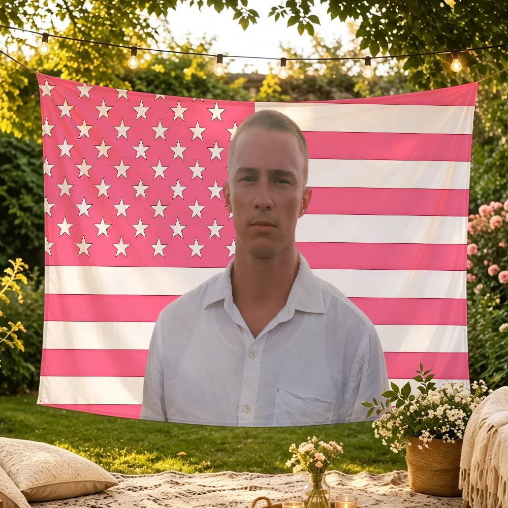 A young man stands in front of a large American flag tapestry featuring pink and white stripes and stars.