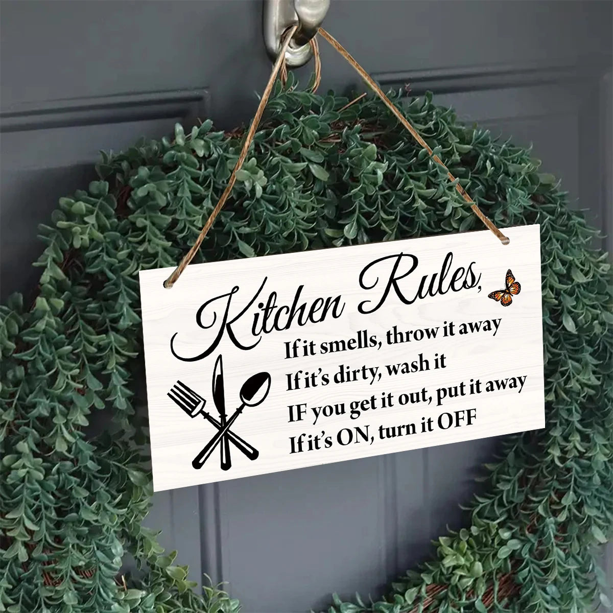 A wooden hanging sign with kitchen rules written on it, displayed on a green faux garland.