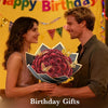 A couple is presenting a bouquet of red glitter artificial roses to each other against a colorful birthday banner.