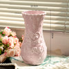 A pink ceramic vase with embossed floral patterns sits on a white cloth near a window.