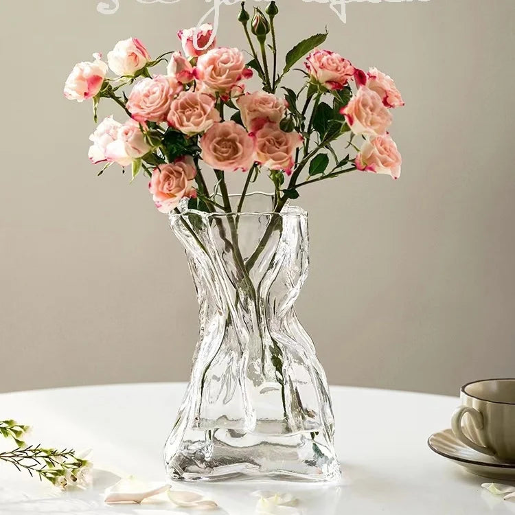 A clear glass vase shaped like a money bag holds pink roses and greenery on a white surface.