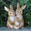 A pair of hand-painted resin rabbit garden statues, one adult and one kit, sitting on a gray surface amidst green foliage.