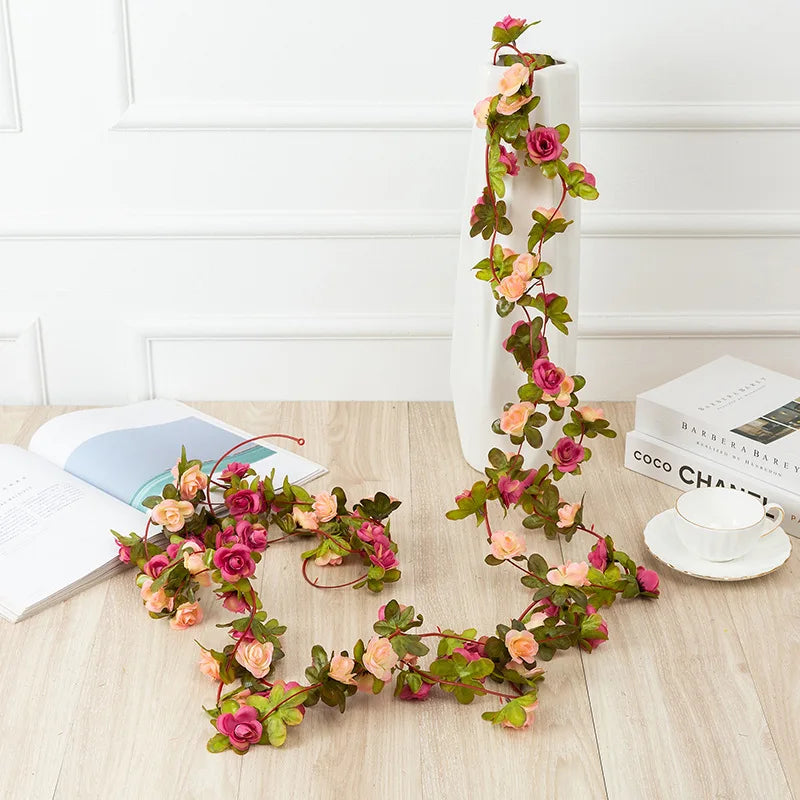 A long silk rose garland with pink and green leaves is displayed vertically in a white vase on a light wooden table.