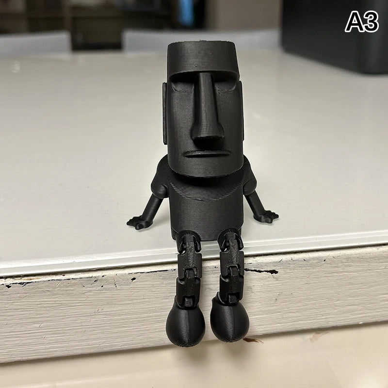 A black 3D printed Moai statue figurine with movable joints standing on a white surface.