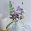 Ramo de flores de ganchillo hecho a mano con flores moradas, blancas y verdes en un jarrón de vidrio transparente.