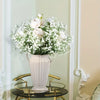 A white artificial baby's breath bouquet in a pale pink vase displayed on a gold-toned table.