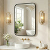 A modern wall mirror with a black metal frame reflects a window and features decorative gold-toned sconces.