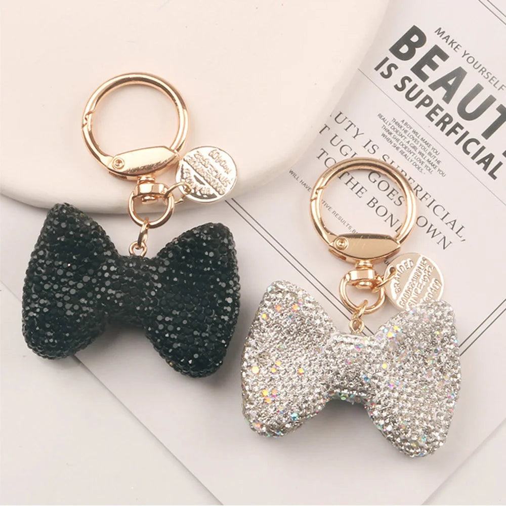 A pair of sparkling rhinestone bow keychains, one black and one silver, displayed on a white card.
