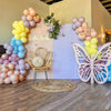 A pastel balloon arch backdrop featuring pink, yellow, and purple balloons, a gold chair, and a large white butterfly decorat