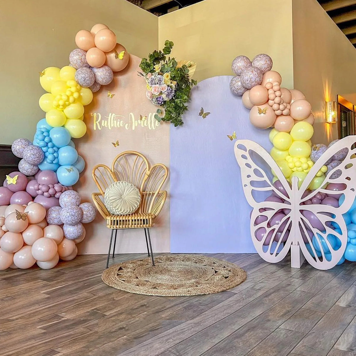 A pastel balloon arch backdrop featuring pink, yellow, and purple balloons, a gold chair, and a large white butterfly decorat