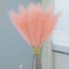 A bouquet of pink pampas grass stems in a gray vase on a white table next to a teacup and a small gold object.