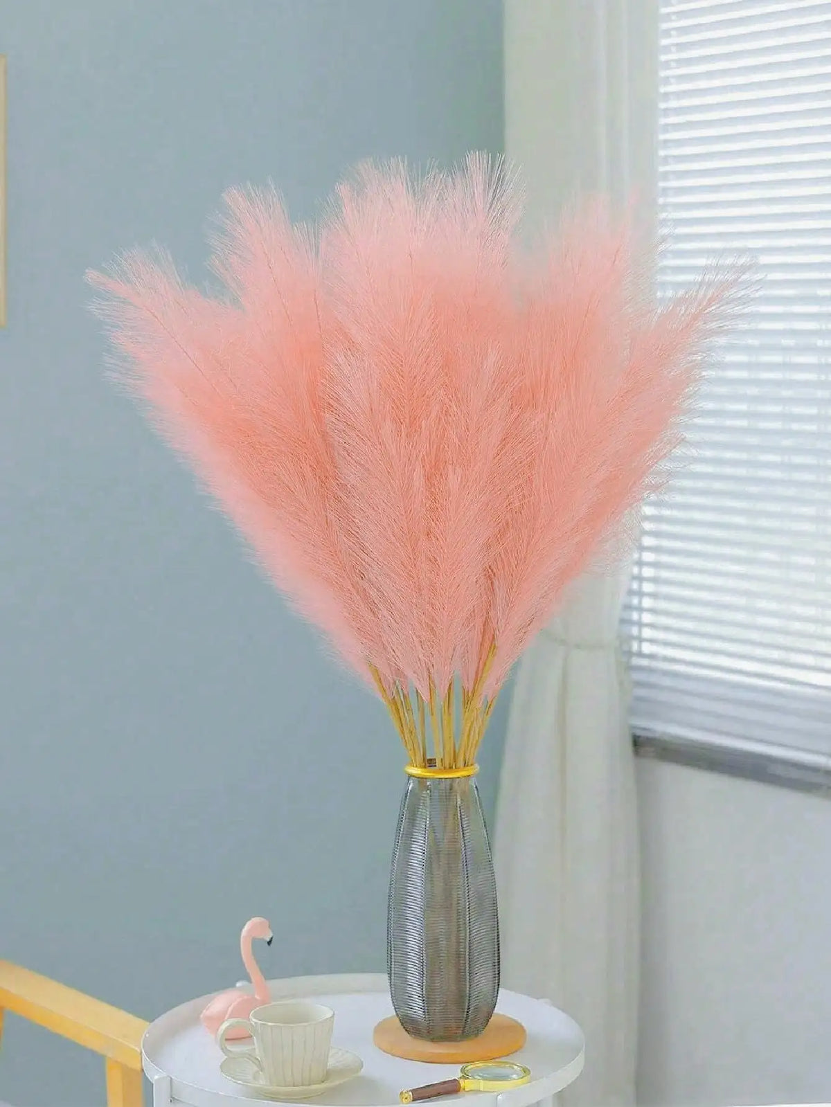 A bouquet of pink pampas grass stems in a gray vase on a white table next to a teacup and a small gold object.