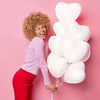 A woman in a pink shirt and red pants is holding a bunch of white heart-shaped latex balloons against a pink background.