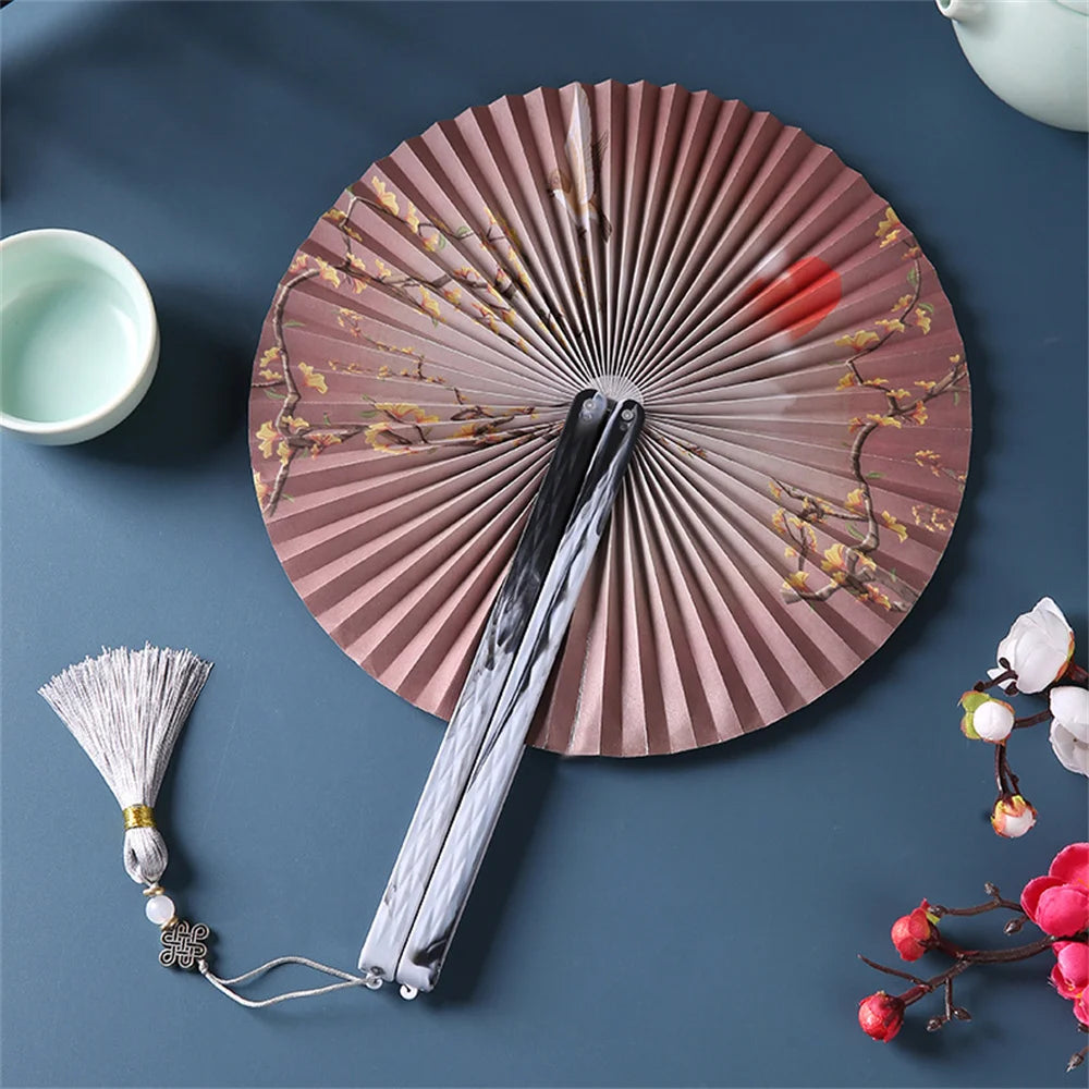 A traditional Chinese folding fan with a pink-brown pleated fan and a white tassel resting on a dark blue surface.