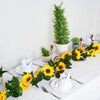 A floral centerpiece featuring yellow sunflowers and green leaves arranged on a white tablecloth with silverware.
