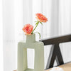 A light green geometric ceramic vase holding a single pink rose in a bright, minimalist setting.