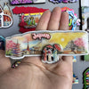 A hand holds a wooden fridge magnet featuring a Japanese cityscape and people against a yellow background.