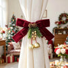 Festive Christmas curtain tie-backs with red bows, pinecones, and gold bells tied on a cream fabric drape.