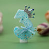 A light blue glittery fabric decoration shaped like a reindeer with a silver crown and gold stars on a white base.