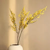 A branch of artificial silk lover grass with yellow flowers in a white ceramic vase against a neutral background.