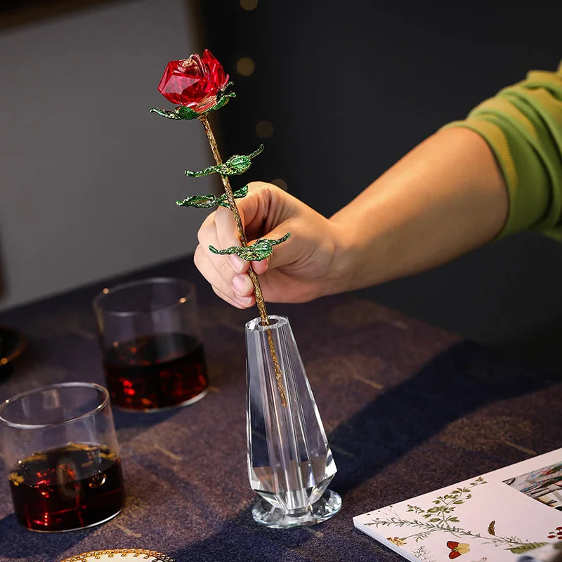 A hand holds a handcrafted crystal glass rose vase with golden leaves on a dark cloth.