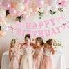 Five girls in pink dresses pose under a glitter pink bow birthday banner decorated with balloons.