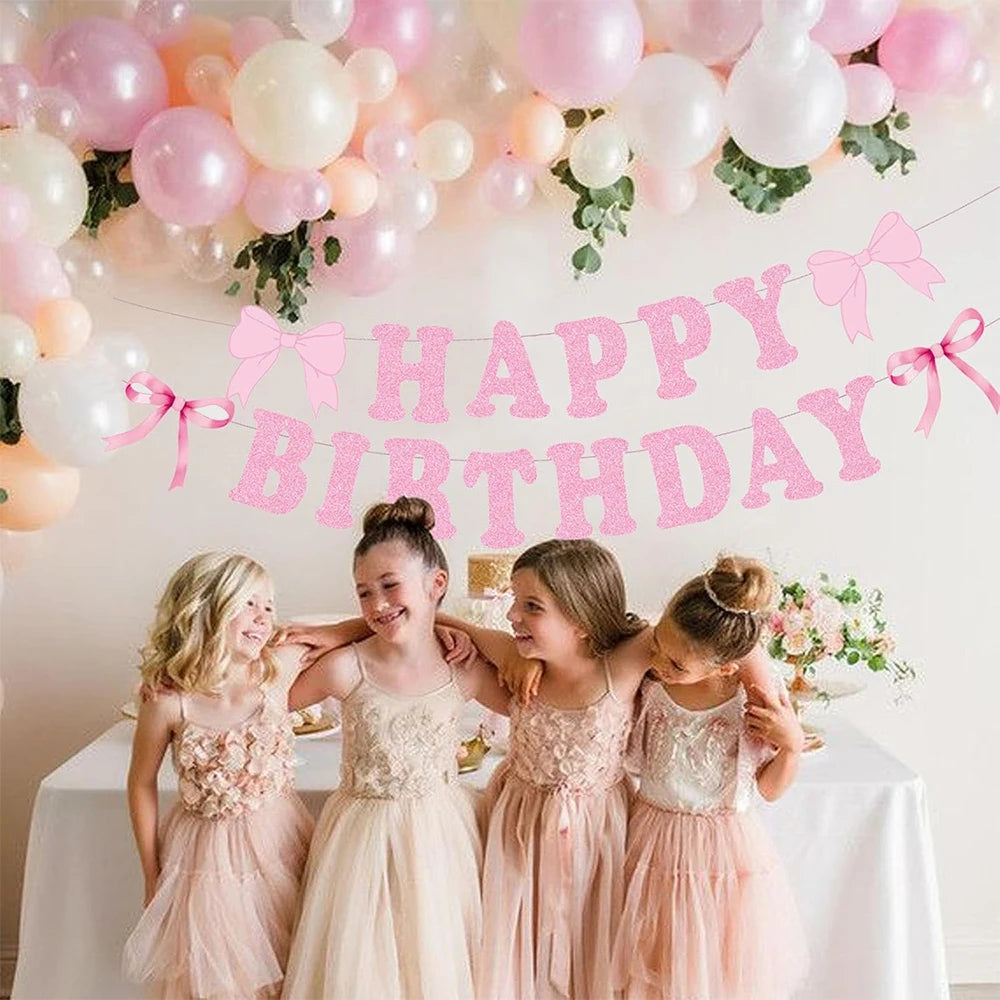 Five girls in pink dresses pose under a glitter pink bow birthday banner decorated with balloons.