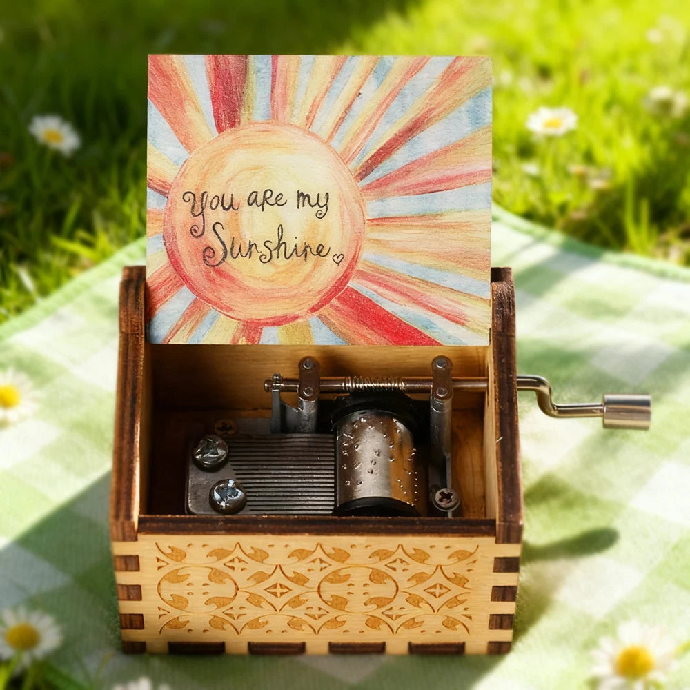 Handbetriebene Holz-Musikbox mit Deckel, der den Text „You are my Sunshine“ trägt, in einer Holzkiste auf grünem Gras.