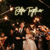 A wedding photo featuring a couple surrounded by people holding sparklers in front of a neon sign that reads Better Together
