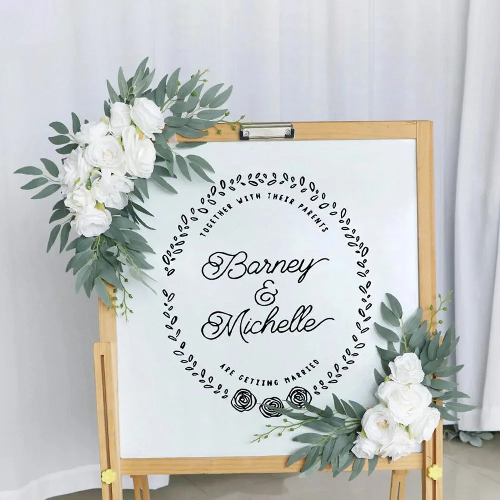 A framed sign featuring a circular wreath design with the text Barney & Michelle are getting married surrounded by white rose