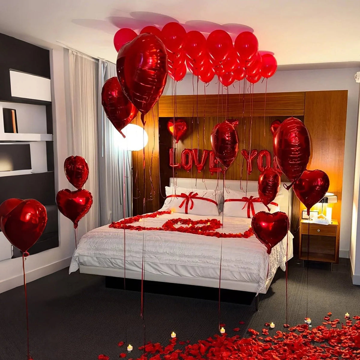A bed decorated with red heart-shaped foil balloons and red rose petals, set in a warmly lit room.